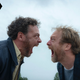Two men in blue jackets scream theatrically into each others' faces, mouths and eyes open wide.
